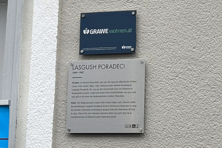 Nahaufnahme einer hell verputzten Hausfassade mit zwei montierten Tafeln: oben eine dunkelblaue Infotafel mit der Aufschrift „GRAWE wohnen.at“, darunter eine silberne Gedenktafel für den albanischen Dichter Lasgush Poradeci (1899–1987) mit biografischem Text in Deutsch und Englisch.