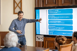 Ein Mann steht neben einem großen Bildschirm und präsentiert eine Folie zum städtebaulichen Rahmenplan mit Prozessschritten wie Flächenwidmung, Bebauungsplanung und Bauverfahren. Er zeigt mit ausgestrecktem Arm auf die Grafik, während mehrere sitzende Zuhörerinnen und Zuhörer im Vordergrund aufmerksam folgen. Der Raum ist holzgetäfelt, auf dem Tisch stehen Unterlagen und ein Glas Wasser.