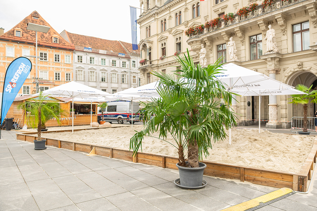 Sandkasten mit Palmen und Polizeiauto vor Gebäuden in Graz